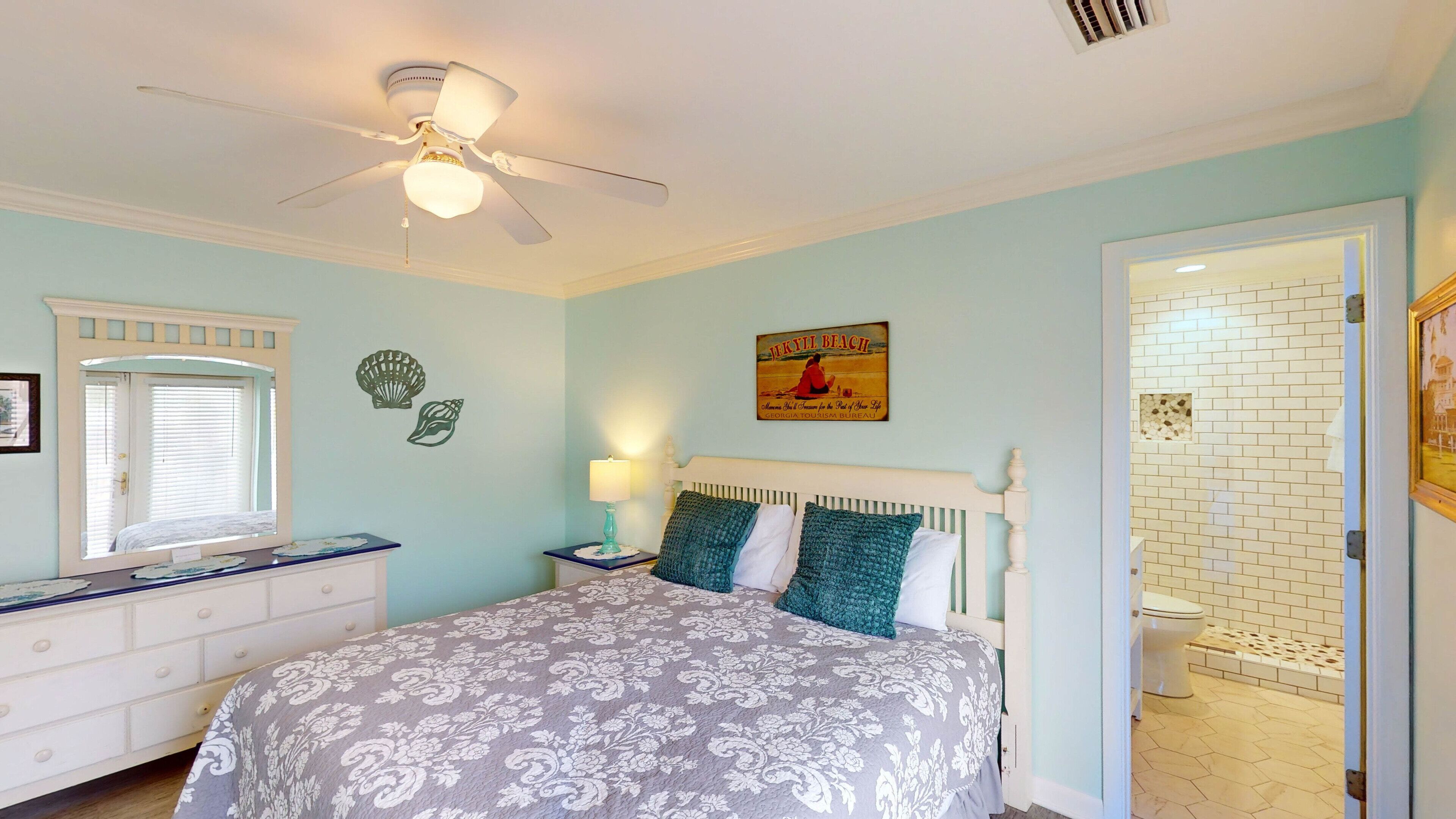 A bedroom with a bed, a nightstand, a ceiling fan, a window with blinds, and a door leading to a bathroom.