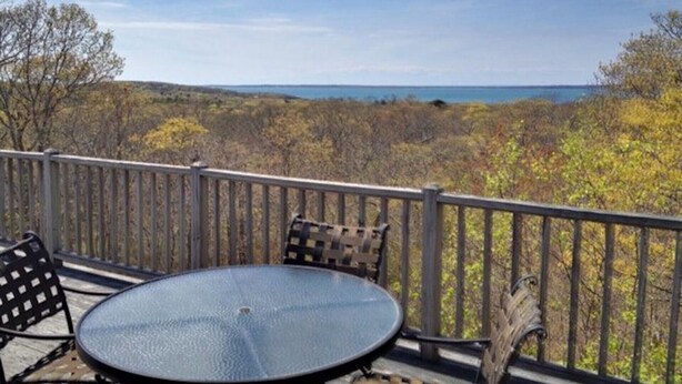Um terraço com uma mesa redonda e cadeiras, com vista para uma área arborizada e um corpo d'água.
