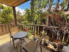 Un patio al aire libre con una mesa redonda y dos sillas, rodeado de exuberante follaje tropical y una cerca de madera.
