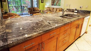 A kitchen with granite countertops, wooden cabinets, a stainless steel sink, and a ceiling fan. - Where Zen Finds You Kihei Garden Condo Split AC Pool Hot Tub Near Beach (Kihei)