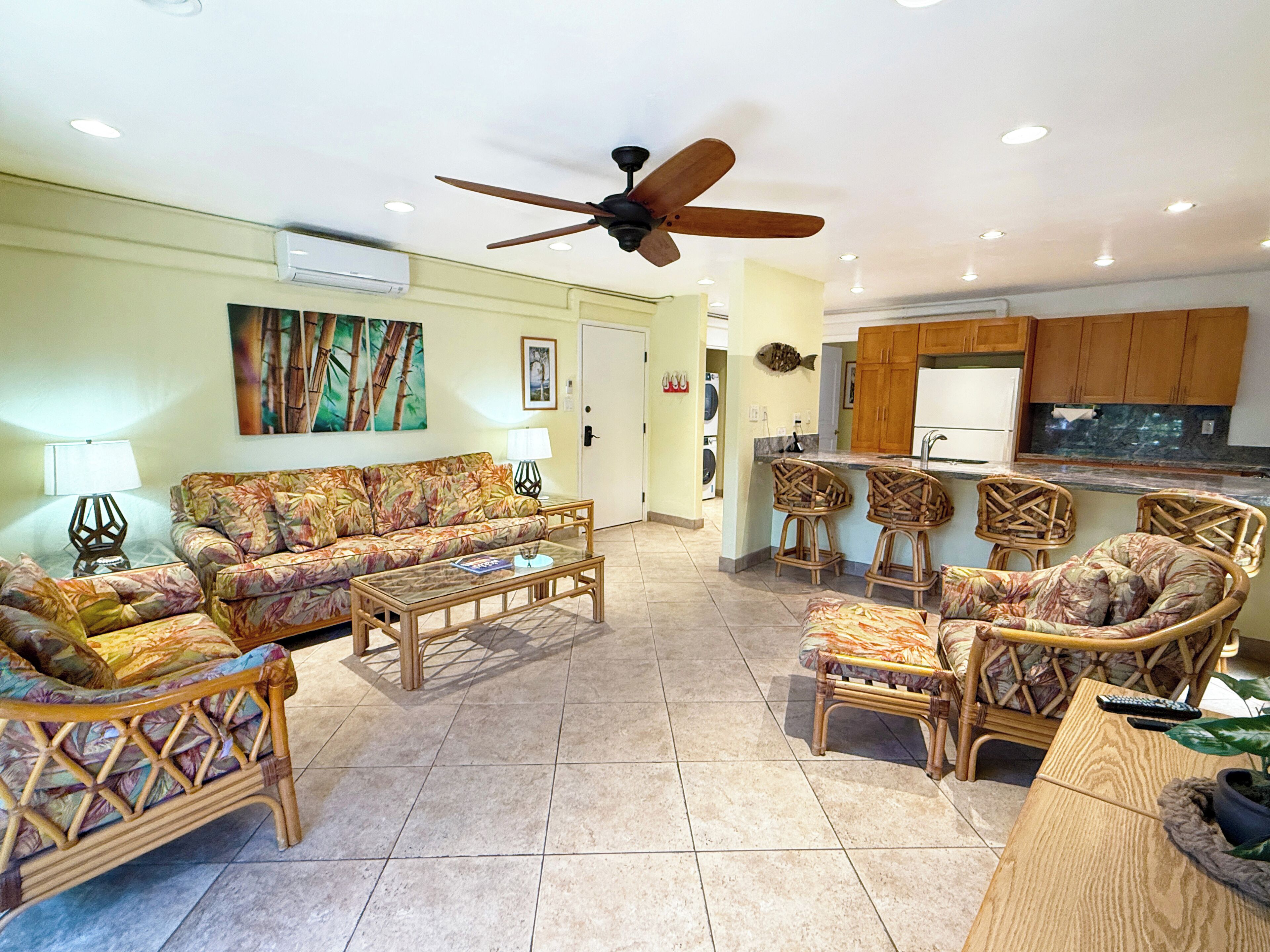 A spacious living area with a sofa, coffee table, and wicker chairs, leading to a kitchen with wooden cabinets and a ceiling fan.