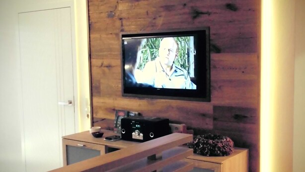 Una sala de estar moderna con un muro decorativo de madera, un televisor de pantalla plana montado sobre un mueble de madera y una pequeña zona de estar con un banco y una mesita auxiliar.