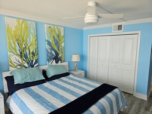 A bedroom with a bed, two wall art pieces, a ceiling fan, a nightstand with a lamp, and a wardrobe.