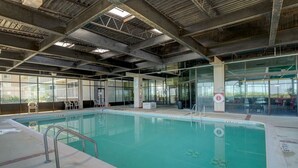 Una piscina interior con paredes de vidrio transparente, barandillas de metal y un techo alto con vigas a la vista.