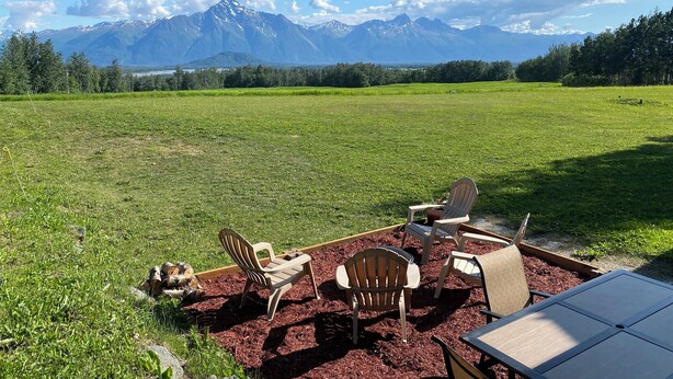 A grassy field with a table and chairs, a wooden chair, and a view of mountains in the background. - Large Home with Outstanding Views & Location (Palmer)