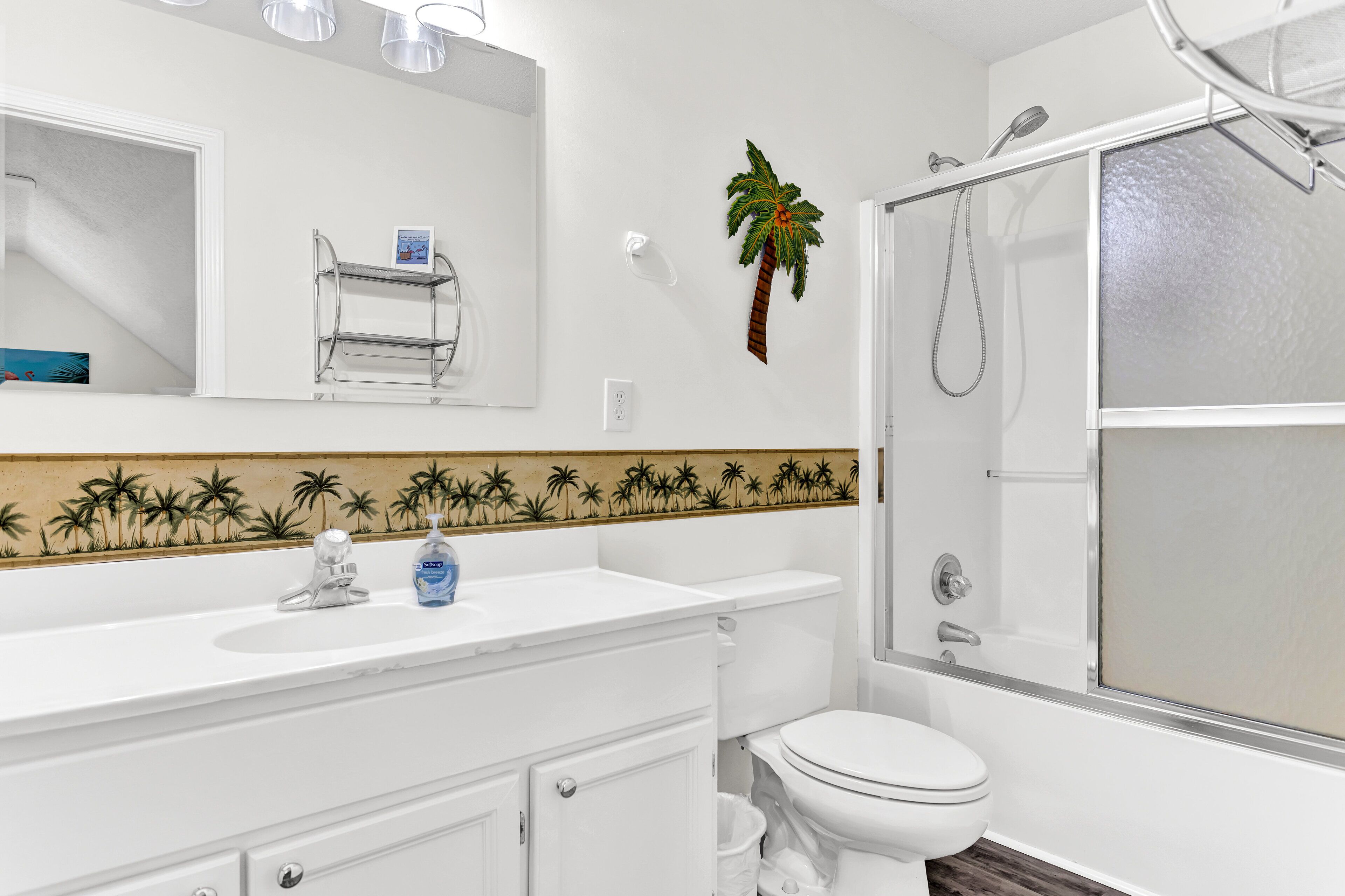 Een badkamer met een wit wastafel, toilet en douche. Boven het wastafel hangt een handdoekhouder en op de muur staat een decoratieve palmboom.