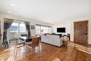 Een moderne woonkamer met een eethoek, houten vloeren, een grijze bank en een flatscreen televisie aan de wand.