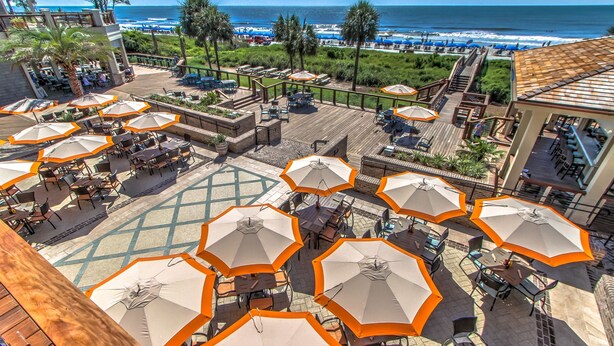 A rooftop patio with multiple tables and chairs, surrounded by orange umbrellas, overlooking a beach and ocean view. - Great Lagoon, Tennis+pool View Townhome W/10min Walk To Golf+Beach+Harbour Town (Hilton Head Island)