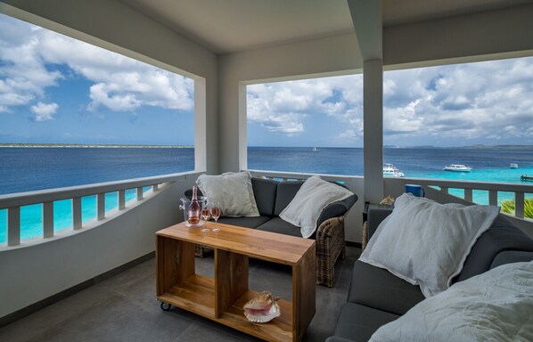 En balkong med utsikt över havet, en soffa, ett träbord med glas och ett snäckskal.