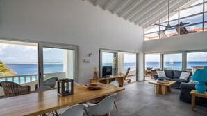 Ein großzügiger Wohnbereich mit einem Holzesstisch, weißen Stühlen, einer Couch und einem Fernseher. Große Fenster bieten Blick aufs Meer.