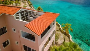 Ein Hotel mit roten Ziegeldächern, Balkonen mit Liegestühlen und Blick auf das türkisblaue Meer.