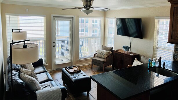 Ein modernes Wohnzimmer mit Sofa, Hocker und Couchtisch. Eine Deckenventilator, ein an der Wand montierter Fernseher und ein Fenster mit Jalousien sind ebenfalls vorhanden.