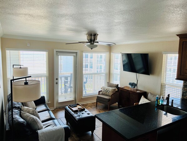 Ein modernes Wohnzimmer mit Sofa, Hocker und Couchtisch. Eine Deckenventilator, ein an der Wand montierter Fernseher und ein Fenster mit Jalousien sind ebenfalls vorhanden.