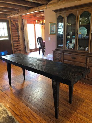 A rustic wooden dining table in a room with a wooden floor, a glass cabinet with dishes, and a chair in the background.