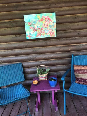 A wooden cabin interior with two blue wicker chairs, a small table with a basket of plants, a bowl, and a painting of birds and flowers on the wall.