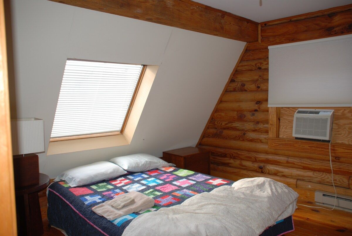 A bedroom with a sloped ceiling, a window, a bed with a quilt, a bedside table, and a wall-mounted air conditioner.