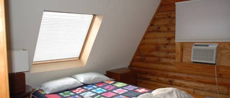 A bedroom with a sloped ceiling, a window, a bed with a quilt, a bedside table, and a wall-mounted air conditioner.