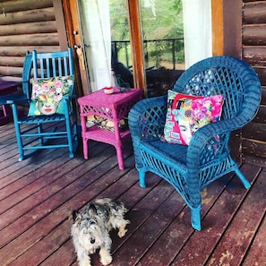 Une terrasse en bois avec une chaise en osier bleue, une table en osier rose et une chaise à bascule bleue agrémentée de coussins à motifs floraux.