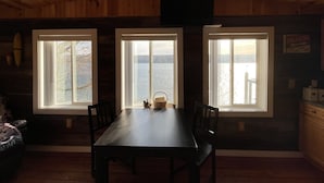 A dining area with a table and chairs, three windows with views of a body of water, and wooden walls. - Unique Space-Great View, East side Sunsets a Plus -2 Nights or More :) (Niles)
