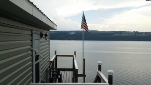 A wooden deck with a railing, a flagpole with an American flag, and a view of a calm lake with distant hills. - Unique Space-Great View, East side Sunsets a Plus -2 Nights or More :) (Niles)