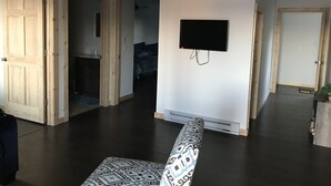 Amplia sala de estar con un televisor de pantalla plana grande montado en la pared, un otoman decorativo en primer plano y piso de madera oscura.