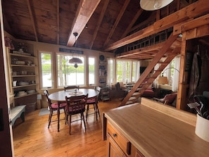 A rustic interior with a wooden dining table, chairs, a staircase, and a view of the outdoors through large windows.