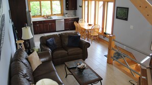 A living room with a brown leather sofa, a coffee table, and a dining area with wooden chairs and a table. There is a kitchen in the background with dark wooden cabinets and a window. - Modern Lakefront home (Hammondsport)