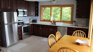 A kitchen with dark wooden cabinets, stainless steel appliances, a large window, and a wooden dining table with chairs. - Modern Lakefront home (Hammondsport)