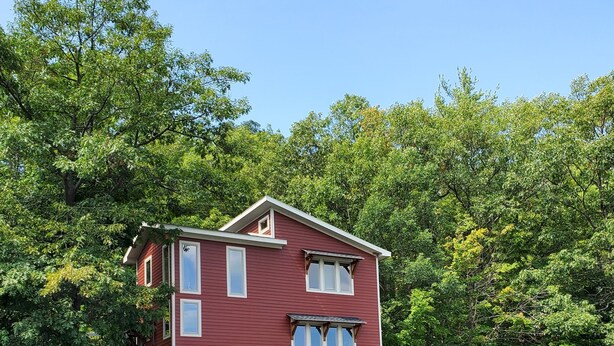 A red two-story house with a dock extending into a body of water, surrounded by dense green trees. - Modern Lakefront home (Hammondsport)