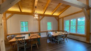 Uma ampla sala de jantar com mesa e cadeiras de madeira, janelas grandes e teto alto com vigas aparentes.