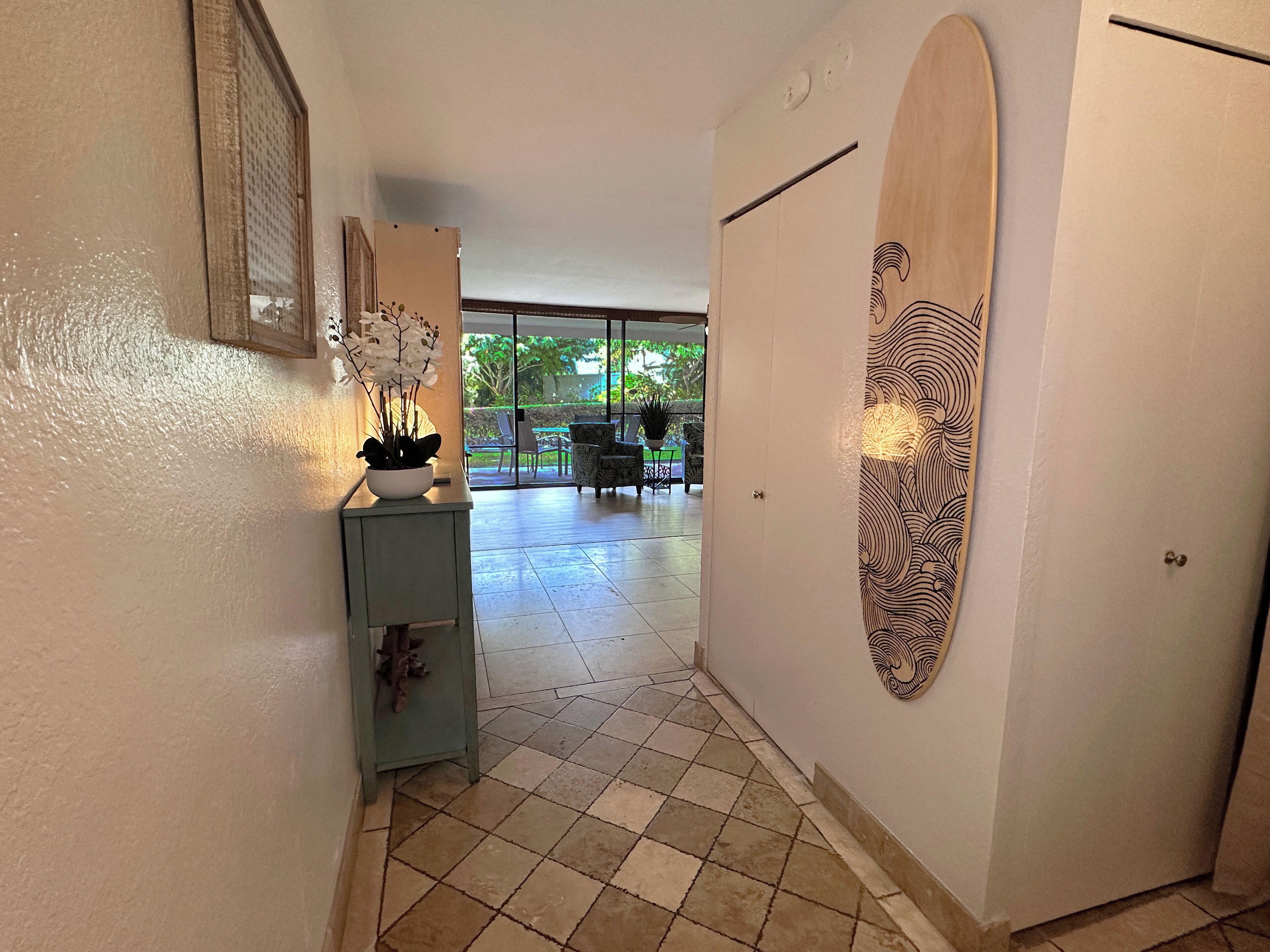 A hallway with a tiled floor, a potted plant, a wooden door with a decorative panel, and a view of an outdoor patio area.