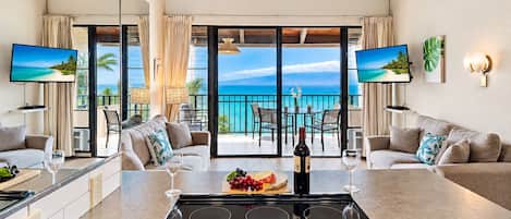 Una cocina moderna con una isla central, un comedor con vistas al océano y una sala de estar con sofá y televisor.