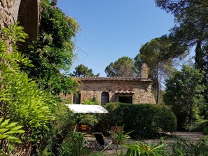 Un bâtiment rustique en pierre, doté d’un toit en tuiles, entouré d’une végétation luxuriante et surplombé d’un ciel bleu clair.