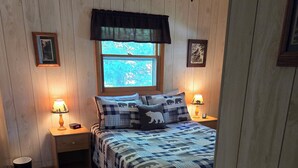 Quarto com uma cama, duas mesas de cabeceira com luminárias, uma janela com vista para árvores e paredes de madeira.