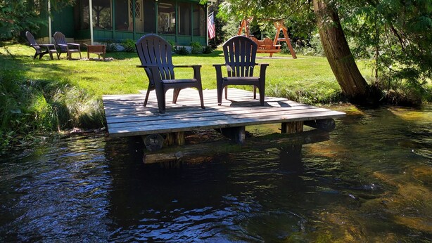 Um cais de madeira com duas cadeiras Adirondack e uma mesa, cercado por uma paisagem verde exuberante e uma cabana ao fundo.