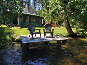 A wooden dock with two Adirondack chairs and a table, surrounded by a lush green landscape and a cabin in the background. - A Perfect Northern Get-Away On The Scenic Big Creek In Luzerne (Luzerne)