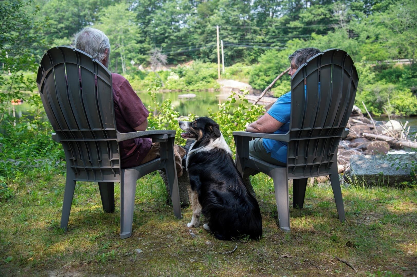 Twee personen zitten tegenover elkaar op stoelen, met een hond ertussen, in een natuurlijke buitenomgeving met begroeiing en een waterpartij op de achtergrond.