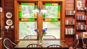A wooden interior with a dining table, chairs, a vase with flowers, a bookshelf, and a window with stained glass.
