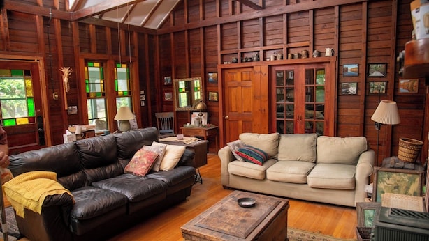 A cozy living room with wooden walls, a black leather sofa, a beige sofa, and a vintage trunk.