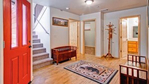 A spacious living room with hardwood floors, a red door, a staircase, a wooden bench, a rug, and a tall wooden stand. - Explorers Retreat (Yosemite National Park)