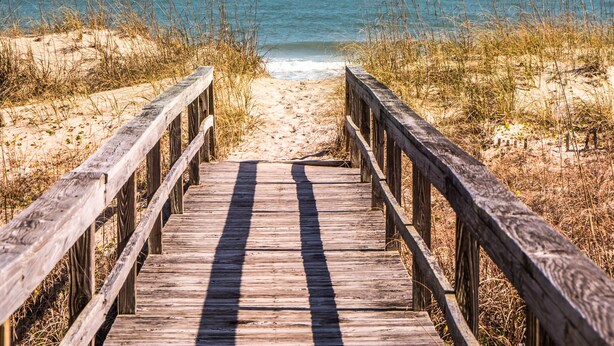 Une promenade en bois qui mène à une plage bordée de sable et d’herbe de chaque côté.