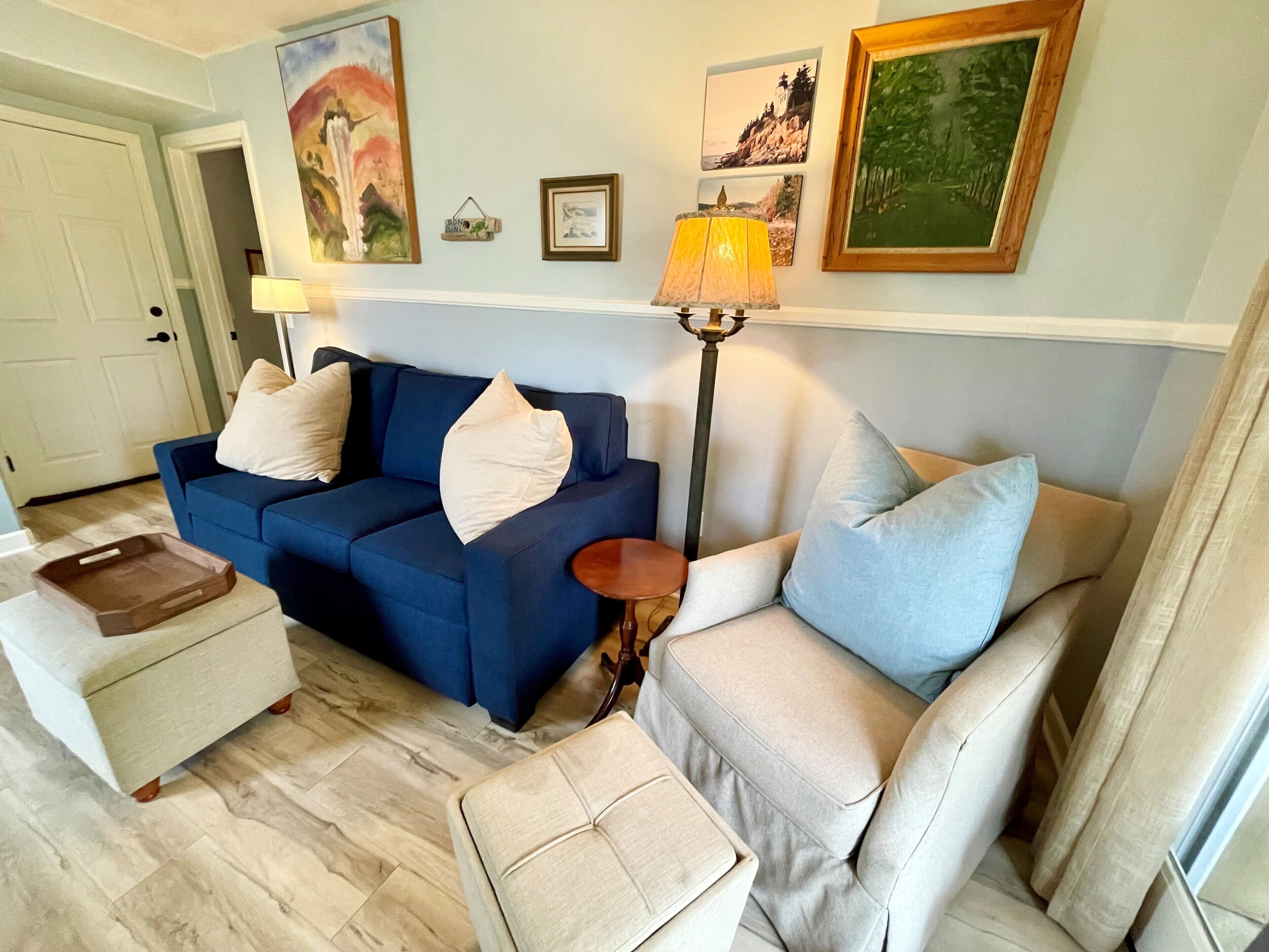 Una sala de estar con un sofá azul, un sillón beige, una mesita y una lámpara de pie. Se observan cuadros enmarcados en la pared y una puerta al fondo.