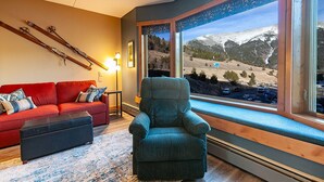 A cozy living room with a red sofa, a teal armchair, and a large window offering a mountain view. -  LUXURY STUDIO+BUNK ROOM, VIEWS, MINS TO LIFTS /SHOPS, ONSITE HOT TUB, GARAGE (Copper Mountain)