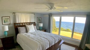A bedroom with a large bed, wooden headboard, two bedside tables with lamps, a ceiling fan, and a view of the sea through large windows. - Prime fall week Oct 10-17 available! Enjoy panoramic ocean views, near Camden (Northport)