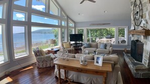 A spacious living room with large windows overlooking a body of water, a ceiling fan, and a stone fireplace. - Prime fall week Oct 10-17 available! Enjoy panoramic ocean views, near Camden (Northport)