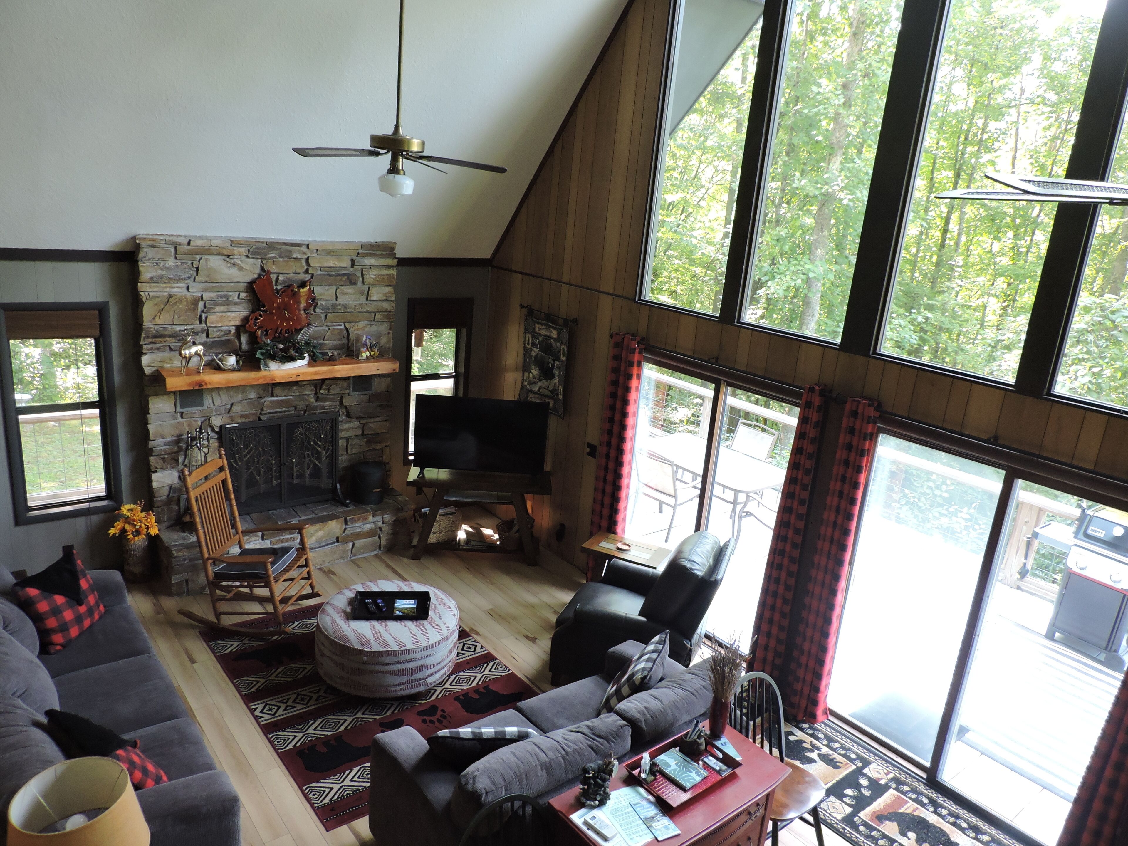 A spacious living room with a stone fireplace, a ceiling fan, and large windows offering a view of the outdoors.
