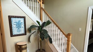 Una escalera con barandillas de madera, una planta en maceta, un cuadro enmarco y un taburete pequeño.