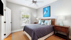 Un dormitorio con cama, mesitas de noche, ventilador de techo, ventana con persianas y una obra de arte colgada en la pared.