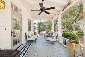 Une véranda couverte avec un ventilateur de plafond, deux chaises, une table et une vue sur une maison au loin.
