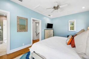 A bedroom with a bed, a television, a ceiling fan, and a door leading to another room.